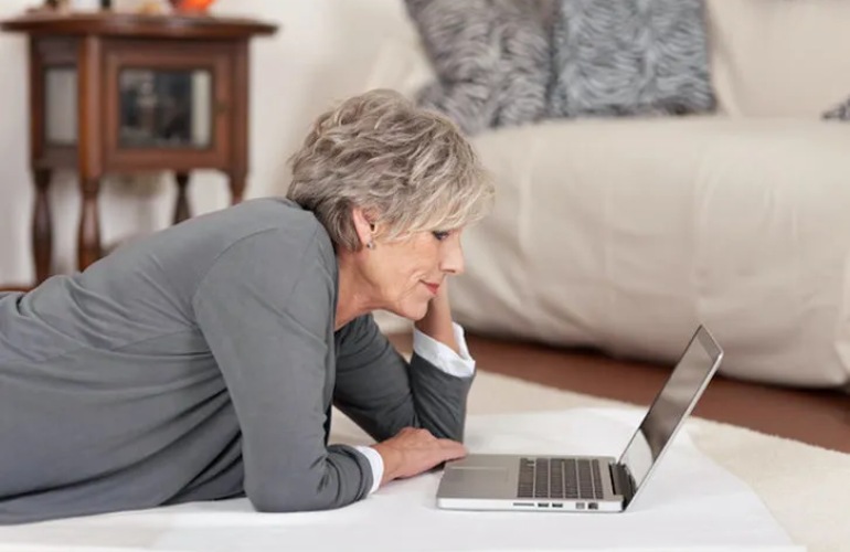 A senior adult woman lays on the floor with a laptop in front of her.