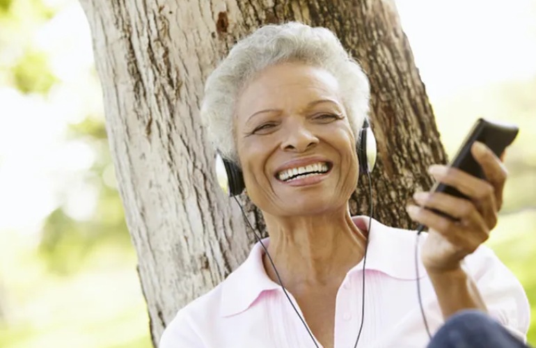 Senior adult woman listening to music while leaning up against a tree.