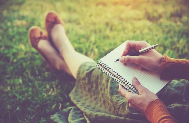 Close up of a woman writing in her journal while laying in the grass.