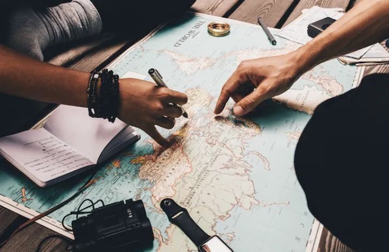 Closeup of a map with people pointing at destinations and writing in a journal.