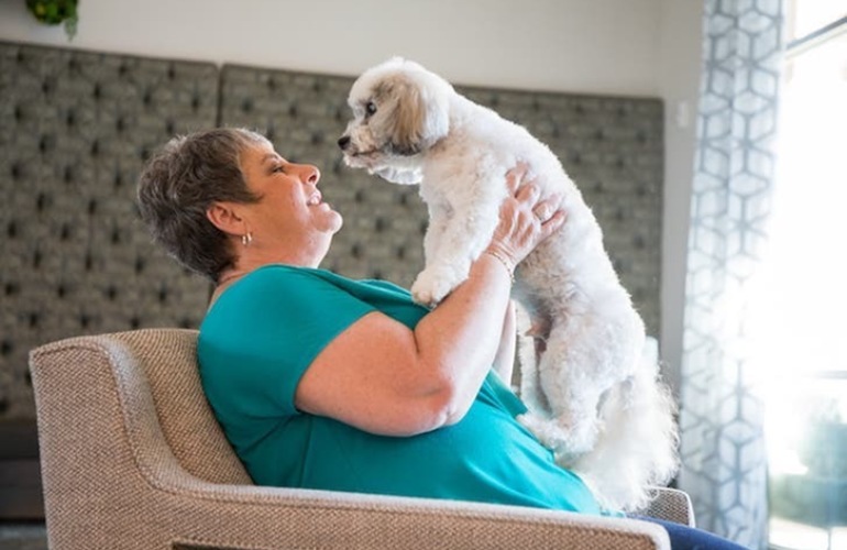 A senior adult woman holds her dog up while seated in a chair.