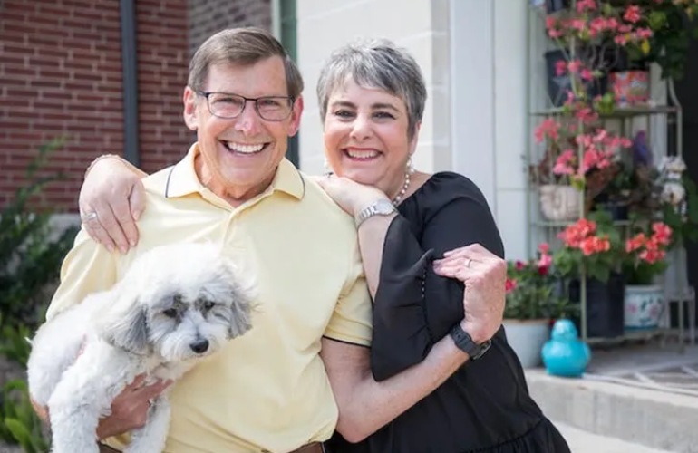 A senior adult couple stand in front of their home holding their pet dog.