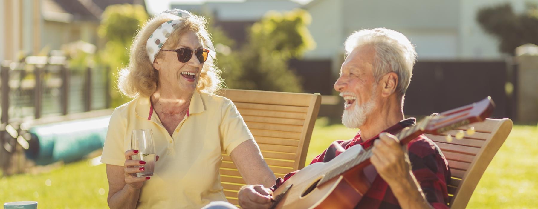 man sits outdoors and plays a guitar for a woman with a drink in her hand