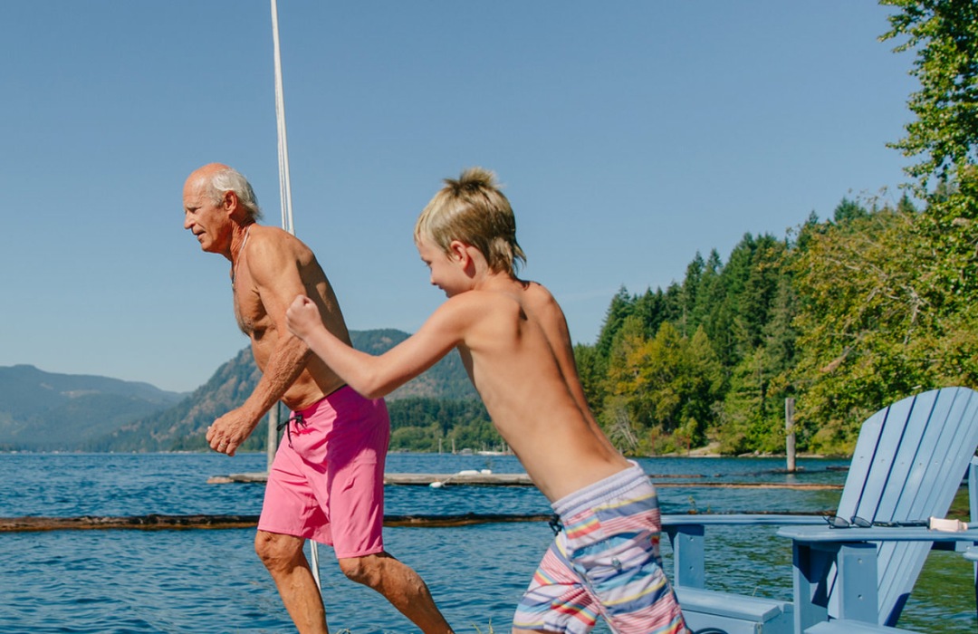 A grandfather and his grandchild run on a dock to jump in a lake.