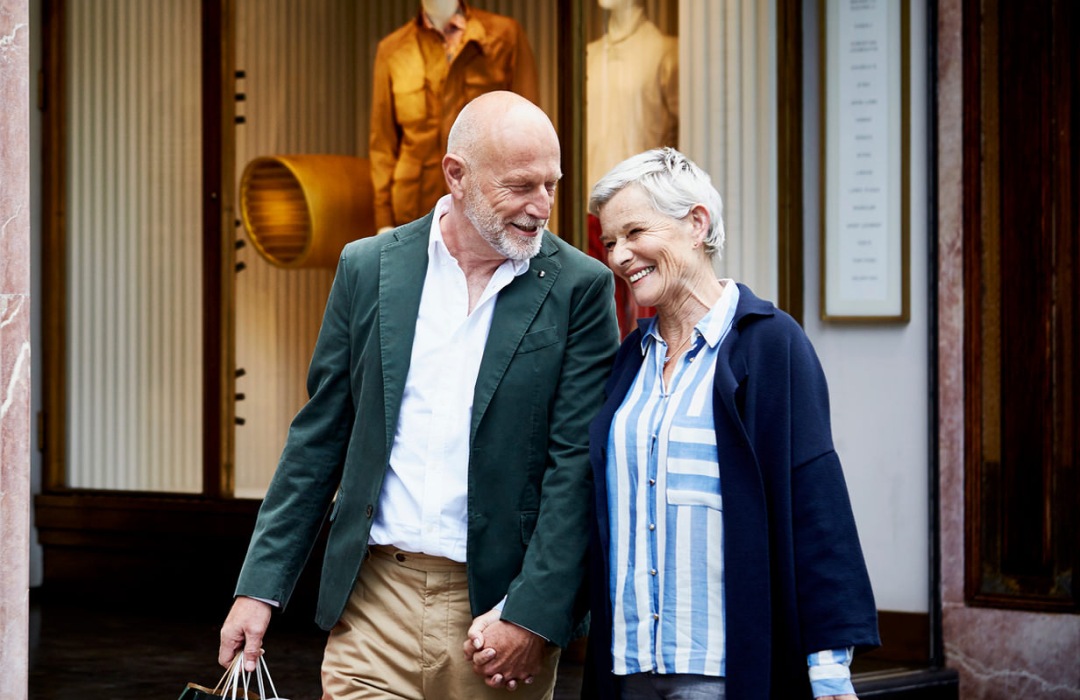 Senior couple walk beside shops in a downtown area holding hands and shopping bags.