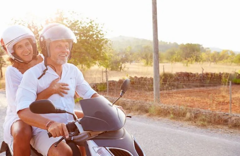 A senior adult couple riding a scooter in the countryside.