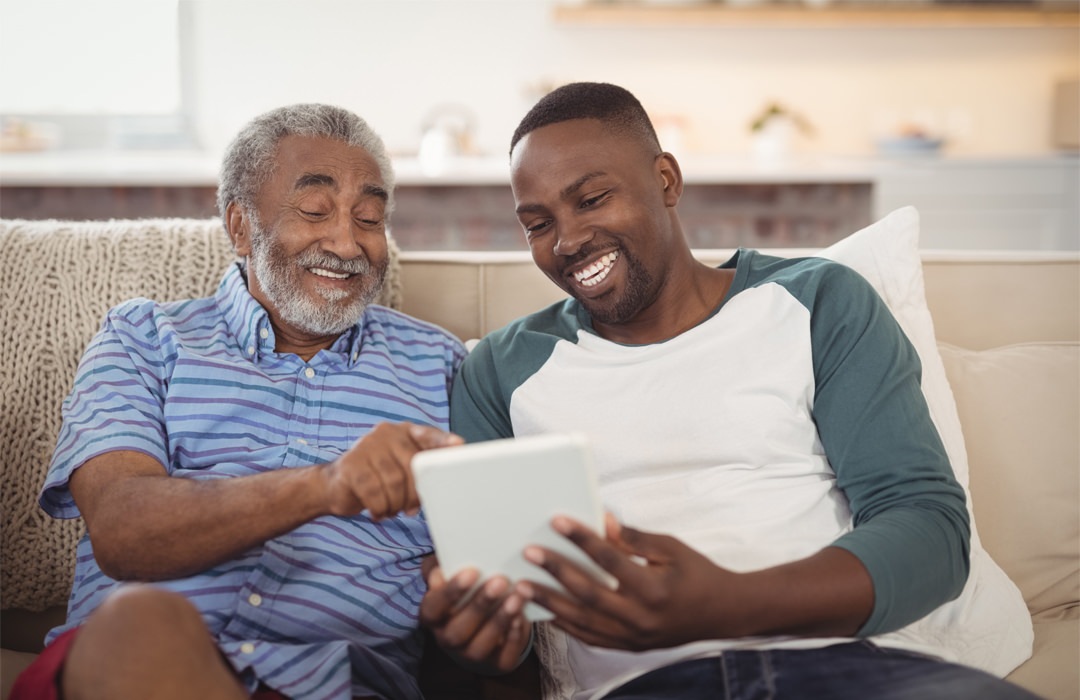 Father and son sitting on a couch in a living room looking at a tablet together.