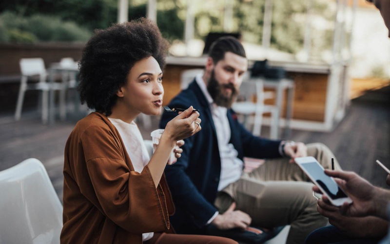 A beautiful community at the heart of all your passions woman sits and eats outside while someone holding a cell talks to her