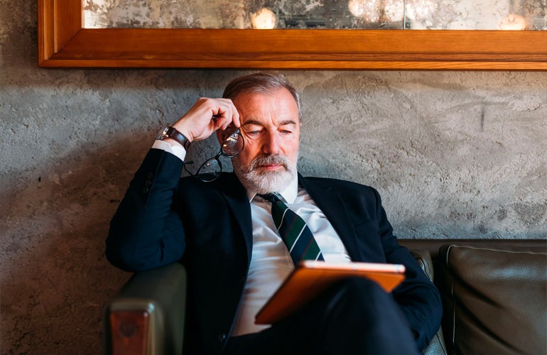 A senior businessman sits on a couch reading his tablet.