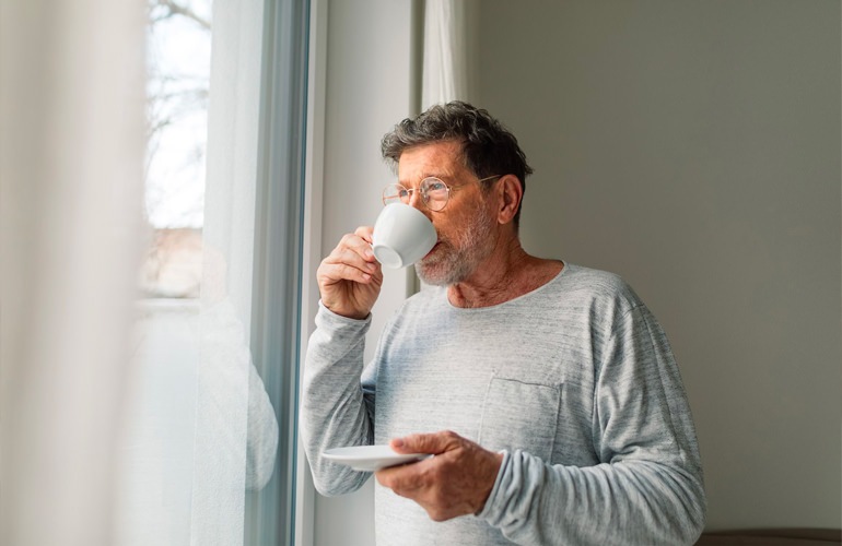 A senior man sips on some coffee while looking out a window in his home.