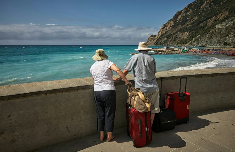 A senior adult couple with traveling baggage looking out at the ocean.